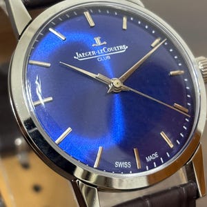 Peut inclure: Gros plan d'une montre-bracelet argentée avec un cadran bleu foncé et un bracelet en cuir marron. Le cadran de la montre présente des aiguilles et des index dorés, avec le texte "JAEGER-LECOULTRE CLUB SWISS MADE".