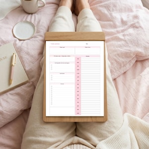 Pu&ograve; includere: Un foglio di pianificazione giornaliera su una cartellina in legno con sfondo bianco e intestazioni e linee rosa. Il testo include Daily planner, Date, Today's goal, Today's focus, 10 positive tasks, Schedule, Top Priorities e To do list.