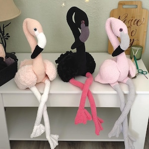 Puede incluir: Tres peluches de flamencos hechos a mano con ganchillo en rosa, negro y rosa claro. Cada flamenco tiene un cuello largo, un pico negro y patas largas. El flamenco de la izquierda tiene patas blancas, el del medio tiene patas rosas y el de la derecha tiene patas grises.