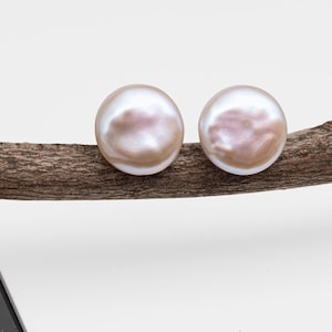 May include: Two large, round, pearl earrings with a lustrous, iridescent surface. The earrings are a soft, creamy color with subtle pink and peach hues. They rest on a textured, brown wooden branch against a white background.