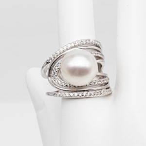May include: A silver ring with a large, round, white pearl at its centre. The ring has a swirling design with multiple bands encrusted with small, clear gemstones. The ring is displayed on a white surface.