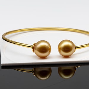 May include: A gold-colored bangle bracelet with two large, round, golden-brown pearls at each end. The bracelet is set against a white and black background, with a reflection of the bracelet visible.