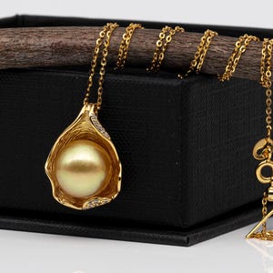 Könnte beinhalten: Eine goldene Anhängerkette mit einer großen, runden, goldenen Perle, die in einer filigranen Goldfassung gefasst ist. Der Anhänger hängt an einer zarten Goldkette. Die Halskette liegt auf einer schwarzen Oberfläche.