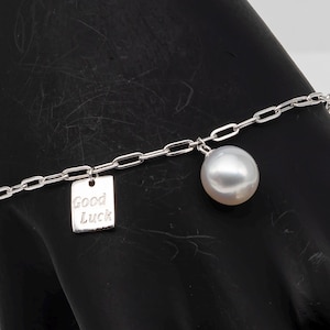 May include: A silver chain bracelet with a large, round, white pearl charm and a small rectangular charm that reads "Good Luck". The bracelet is displayed against a black background.