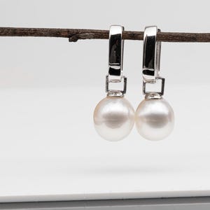 May include: A pair of pearl drop earrings. Each earring has a large, lustrous, white pearl suspended from a silver-coloured, hinged hoop. The earrings are displayed against a white background, hanging from a thin, brown twig.