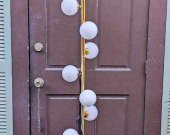 May include: A tall floor lamp featuring a vertical gold-toned metal pole with nine spherical, frosted white glass globes attached at varying heights along the stem. A black power cord hangs down the side of the central pole.