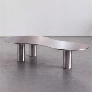 Könnte beinhalten: Ein niedriger Couchtisch mit einer Oberfläche aus gebürstetem silberfarbenem Metall. Die Tischplatte hat eine unregelmäßige, organische Nierenform mit glatten, abgerundeten Kanten. Er steht auf drei dicken, zylindrischen Metallbeinen.