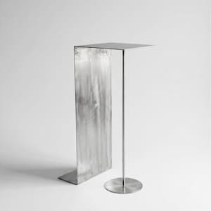 Può includere: Un tavolino in metallo color argento caratterizzato da un piano rettangolare piatto sostenuto da una sottile asta verticale fissata a una base circolare, abbinato a un pannello metallico rettangolare verticale alto e largo.