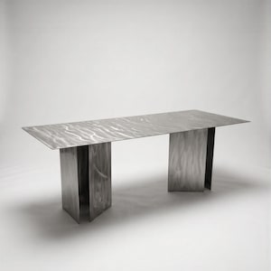 Op de afbeelding: Een rechthoekige eettafel gemaakt van geborsteld metaal met een opvallende golvende, geribbelde textuur over het oppervlak. De tafel heeft twee brede, verticale plaatvormige poten die het lange, platte blad ondersteunen, wat zorgt voor een strakke, industriële uitstraling.