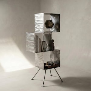 May include: A three-tiered metal shelving unit with a brushed silver finish and four thin, angled legs. The structure consists of three stacked, open-faced cube compartments containing books, a geometric sculpture, a vase, and a camera.