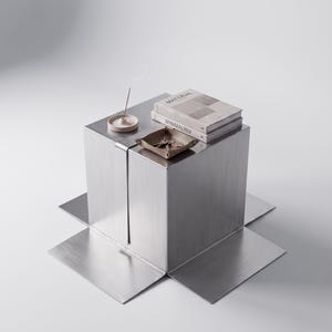 May include: A brushed metal cube table with four flat base extensions. On top sits a beige incense holder with a smoking stick, a beige leather tray containing metal keys, and two stacked books titled Material and Minimalism.