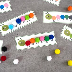 May include: Educational cards featuring cartoon caterpillars with colorful pom-pom bodies. Each card has a green caterpillar head and a series of pom-poms in various colors, including red, orange, pink, and blue. Several loose pom-poms are scattered around the cards.