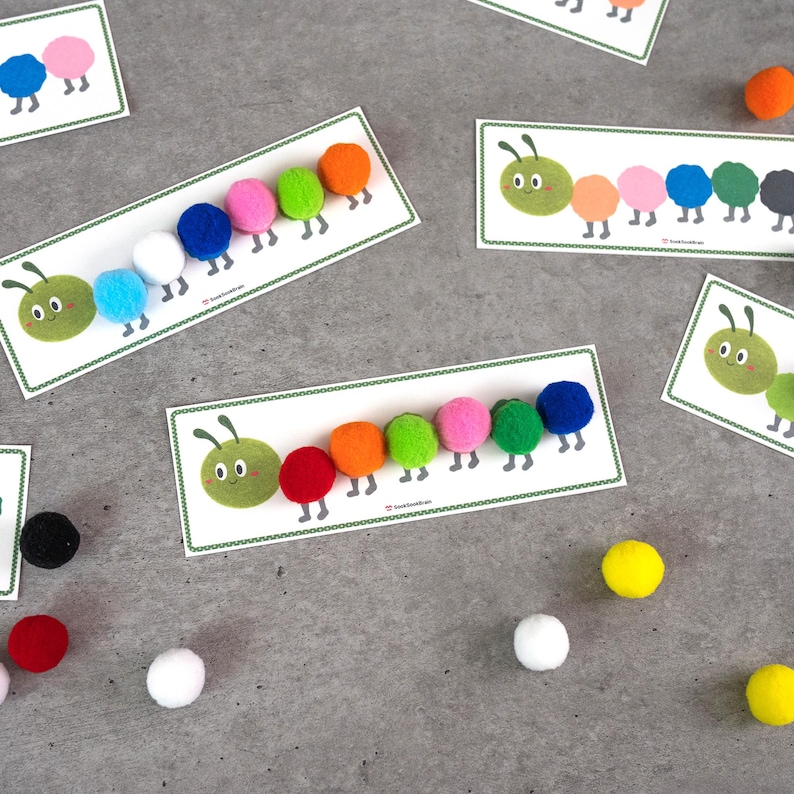 May include: Educational cards featuring cartoon caterpillars with colorful pom-pom bodies. Each card has a green caterpillar head and a series of pom-poms in various colors, including red, orange, pink, and blue. Several loose pom-poms are scattered around the cards.