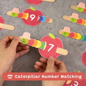 May include: Caterpillar Number Matching game featuring red apple cards with white numbers and colorful caterpillar pieces. The caterpillars have smiling faces and wear pink hats. The game is designed for educational purposes.
