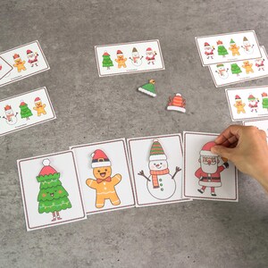 Puede incluir: Tarjetas navide&ntilde;as con ilustraciones de un &aacute;rbol de Navidad, un hombre de jengibre, un mu&ntilde;eco de nieve y Pap&aacute; Noel. Las tarjetas est&aacute;n sobre una superficie gris, con una mano colocando una de ellas. Medidas m&eacute;tricas.