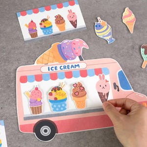 May include: A die-cut paper ice cream truck display featuring a pink body, blue awning, and the text ICE CREAM. It includes various paper ice cream cone and cupcake cutouts with colorful designs, including a pink rabbit-shaped ice cream cone.