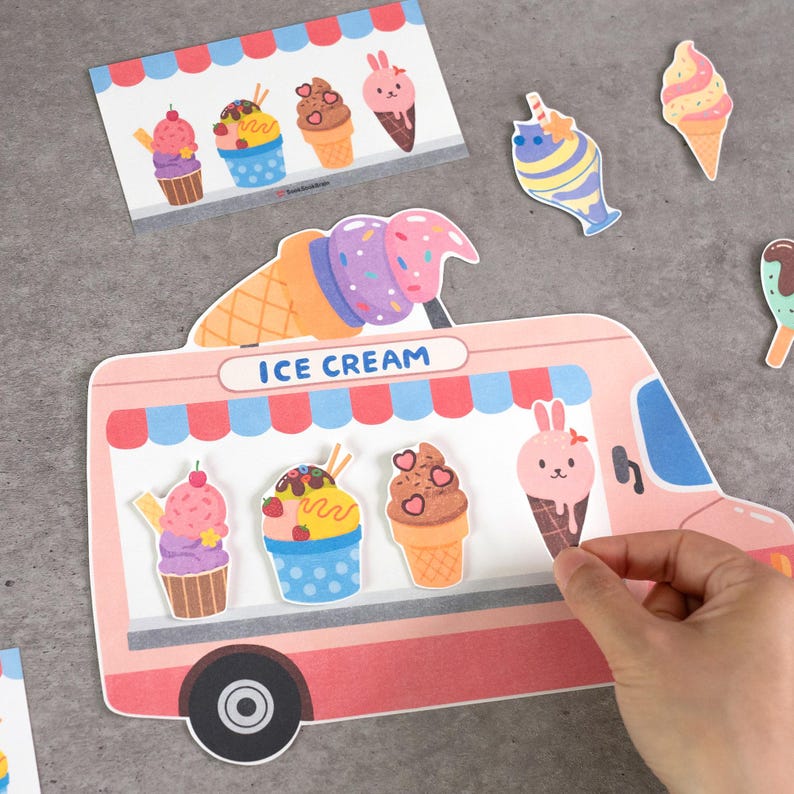 May include: A die-cut paper ice cream truck display featuring a pink body, blue awning, and the text ICE CREAM. It includes various paper ice cream cone and cupcake cutouts with colorful designs, including a pink rabbit-shaped ice cream cone.