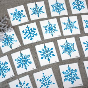 May include: A collection of white cards with blue snowflake designs. Each card is cut in half, creating a puzzle. The snowflakes vary in design, with intricate details. The cards are arranged on a gray surface, suggesting a winter or holiday theme.