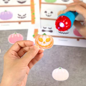 May include: A small, die-cut sticker of an orange jack-o'-lantern with a green stem and yellow facial features. The sticker is being illuminated by a blue and red flashlight, revealing a glowing effect on the pumpkin's surface against a printed sheet.