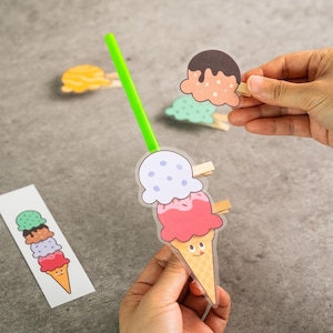 May include: A collection of colorful ice cream cone-shaped paper clips and a bookmark with ice cream illustrations. The paper clips feature various ice cream flavors and are held by small wooden clips. A green pen is also in the image.
