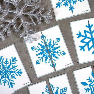 May include: A collection of white cards with blue snowflake designs, some cut in half, arranged on a gray surface. A clear, plastic snowflake decoration is in the upper left corner. The cards have a gray border and the text "SeekBrain" in the bottom right corner.