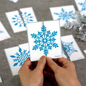 May include: A person is holding a white card with a blue snowflake design, split in two. Other cards with similar snowflake designs are scattered around. The snowflake design is a detailed, symmetrical pattern. The cards have a light gray border.