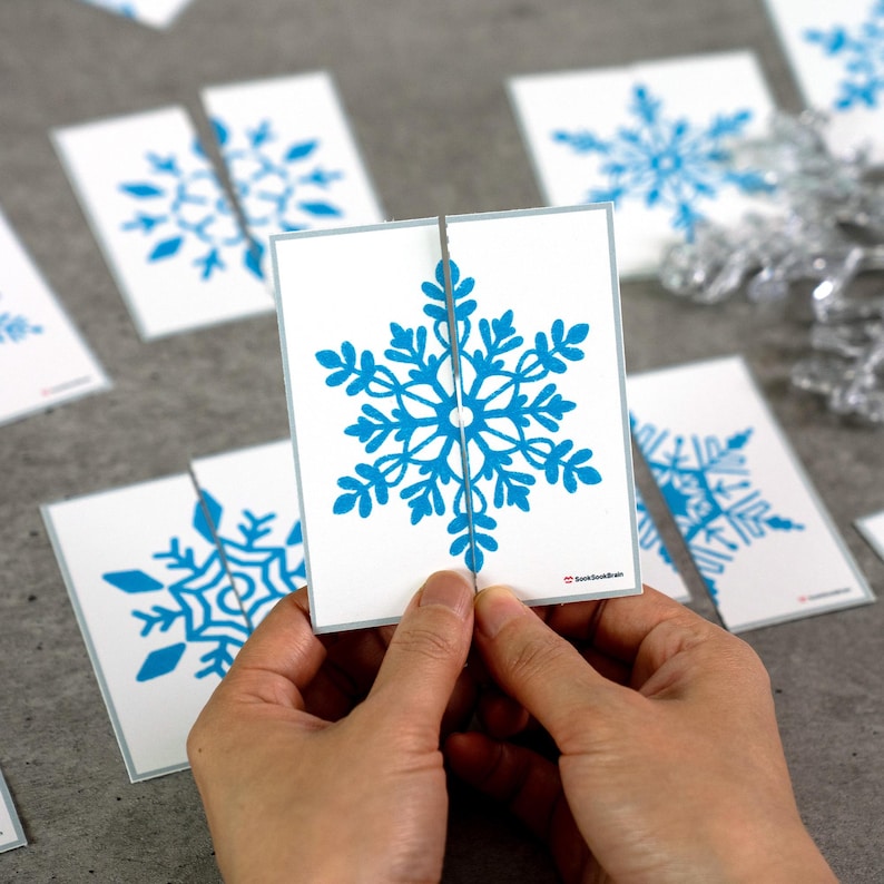 May include: A person is holding a white card with a blue snowflake design, split in two. Other cards with similar snowflake designs are scattered around. The snowflake design is a detailed, symmetrical pattern. The cards have a light gray border.