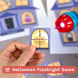 Puede incluir: Un juego de linterna de Halloween con hojas de papel impresas con ilustraciones de casas y ventanas. Una pequeña tarjeta cuadrada muestra una calabaza amarilla en una ventana, mientras una linterna azul y roja ilumina la escena.