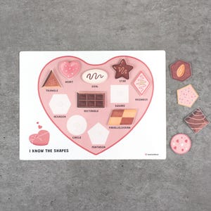 May include: A wooden shape puzzle with a heart-shaped cutout and various geometric shapes. The puzzle includes shapes like triangles, hearts, and stars, with the text "I KNOW THE SHAPES" at the bottom. Additional shapes are placed to the right.