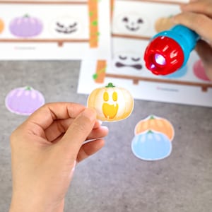 May include: A small, die-cut paper pumpkin cutout with a yellow jack-o'-lantern face, held in front of a printed activity sheet featuring various pumpkin illustrations and a blue and red plastic flashlight emitting a bright white light beam.