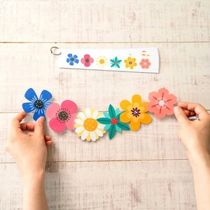 May include: A colorful floral garland with various flower designs in blue, pink, yellow, and green. The garland is held by two hands against a light wood background. A matching floral design is on a white strip.