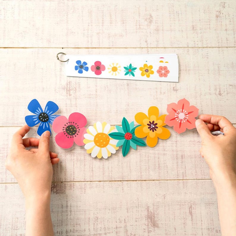 May include: A colorful floral garland with various flower designs in blue, pink, yellow, and green. The garland is held by two hands against a light wood background. A matching floral design is on a white strip.