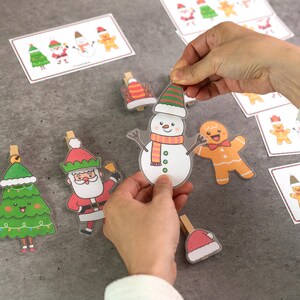 Puede incluir: Una colecci&oacute;n de recortes de papel con tem&aacute;tica navide&ntilde;a, que incluyen un mu&ntilde;eco de nieve, un hombre de jengibre, Pap&aacute; Noel y un &aacute;rbol de Navidad, cada uno adjunto a una pinza de madera. Tarjetas adicionales con dise&ntilde;os similares est&aacute;n dispersas. Los dise&ntilde;os presentan colores brillantes e ilustraciones de estilo caricatura.