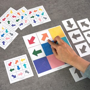 May include: A collection of colorful arrow cards and a larger game board with colored squares. The arrow cards feature various colored arrows pointing in different directions. A hand is pointing at an orange arrow on the game board. Educational game.