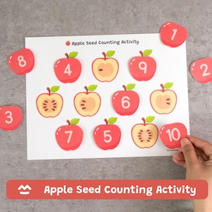 May include: An Apple Seed Counting Activity worksheet featuring a grid of red apple illustrations, some whole and some halved, with numbers printed on them. Several loose red apple-shaped cutouts with numbers are placed around the main sheet.