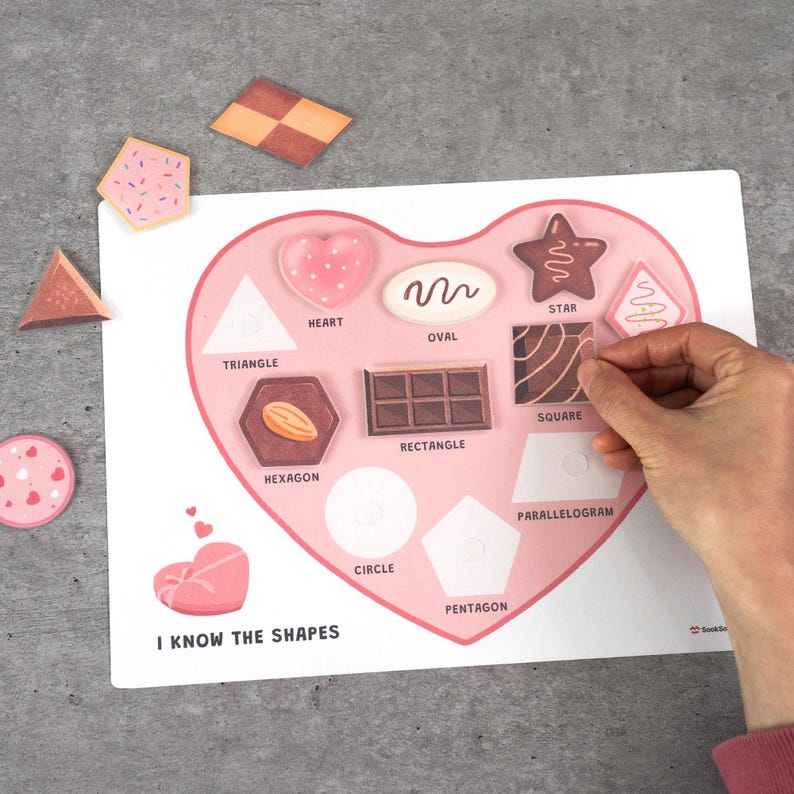 May include: A pink heart-shaped shape matching puzzle with various shapes and corresponding chocolate-themed pieces. The puzzle includes shapes like a heart, star, and oval. The text "I KNOW THE SHAPES" is visible.