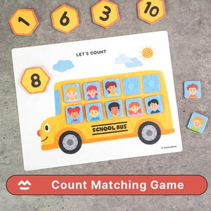 Peut inclure: Jeu d'association de comptage avec un bus scolaire jaune et des visages d'élèves dans les fenêtres. Le jeu comprend des tuiles numérotées et des tuiles d'élèves. Le texte "LET'S COUNT" et "SCHOOL BUS" sont visibles. Conçu pour l'apprentissage des chiffres.