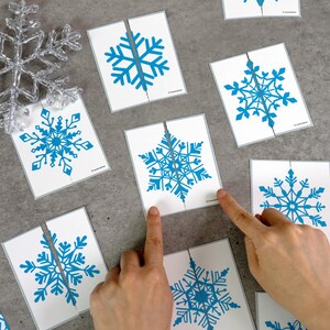 May include: White cards with blue snowflake designs, each cut in half, are arranged on a gray surface. A clear, decorative snowflake is in the upper left corner. Fingers are pointing at the cards, suggesting a puzzle or game.