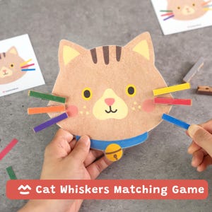 Pode incluir: Um recorte de papelão em forma de gato com rosto bronzeado, listras marrons na testa e uma coleira azul com um sino amarelo. O gato tem seis bigodes de prendedores de roupa coloridos fixados nas laterais em verde, laranja, roxo, amarelo, vermelho e azul.