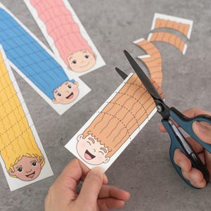 May include: A person is cutting a brown-haired cartoon character with scissors. The character has a smiling face and is part of a paper craft project. Other characters with pink, blue, and yellow hair are visible.