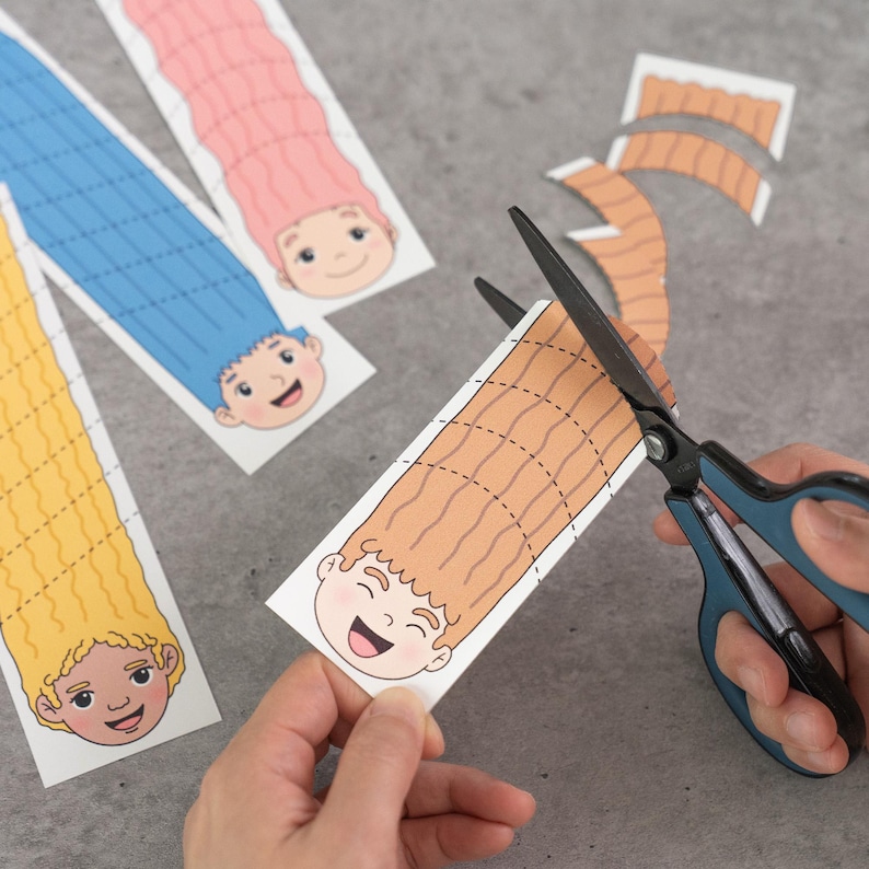 May include: A person is cutting a brown-haired cartoon character with scissors. The character has a smiling face and is part of a paper craft project. Other characters with pink, blue, and yellow hair are visible.