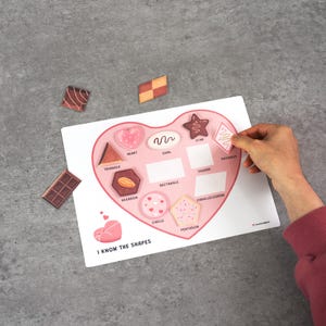 May include: A heart-shaped educational game board with shape cutouts and the text "I KNOW THE SHAPES". The board features various shapes like triangles, stars, and circles. Several shape cutouts are scattered around the board.