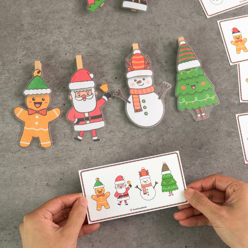 Puede incluir: Decoraciones navide&ntilde;as con un hombre de jengibre, Pap&aacute; Noel, un mu&ntilde;eco de nieve y un &aacute;rbol de Navidad, cada uno sujeto a una pinza de madera. Una persona sostiene una tarjeta con los mismos personajes. Los personajes son de estilo caricatura.