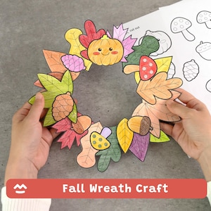 Puede incluir: Una corona de papel artesanal con ilustraciones a color de hojas de otoño, una calabaza sonriente, setas y piñas. La corona es circular con una abertura central. Se ve una hoja de papel separada con dibujos de líneas en blanco y negro.