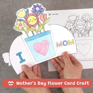 Könnte beinhalten: Bastelarbeit für eine Muttertags-Blumenkarte. Die Karte zeigt einen blauen Blumentopf mit einem rosa Herzen und bunten Blumen. Die Wörter "I" und "MOM" sind sichtbar. Das Bild enthält auch eine gedruckte Vorlage.