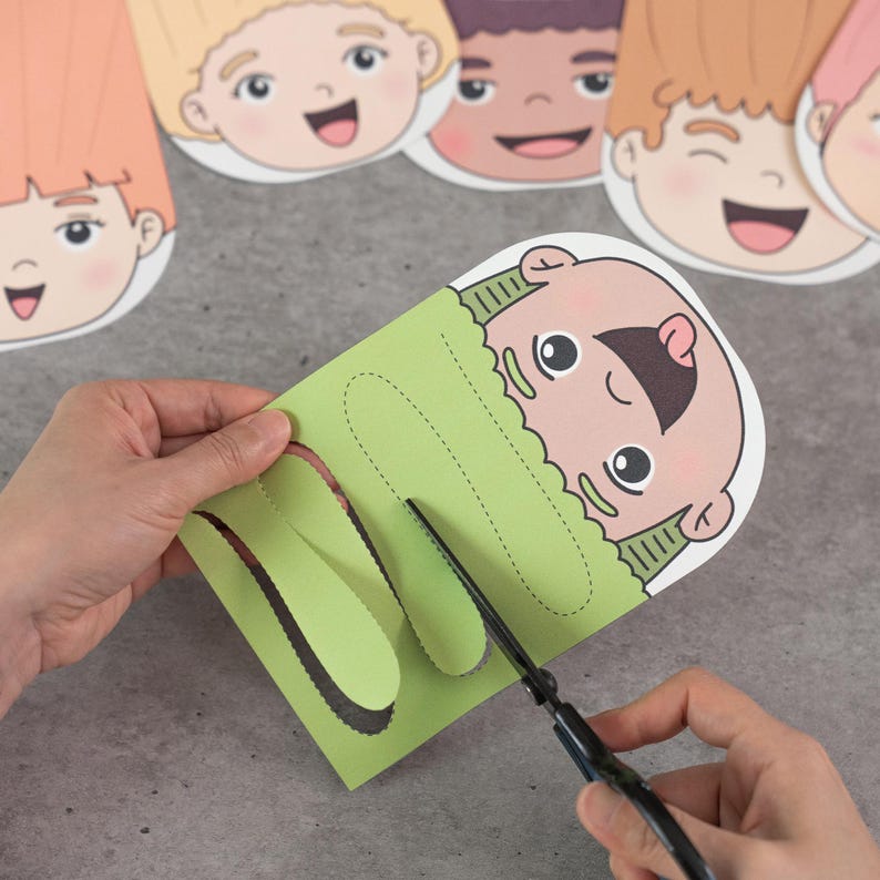 May include: A person is using scissors to cut a green paper craft featuring a cartoon face. Several other cartoon faces are visible in the background. The craft has a cut-out design for a child's face.