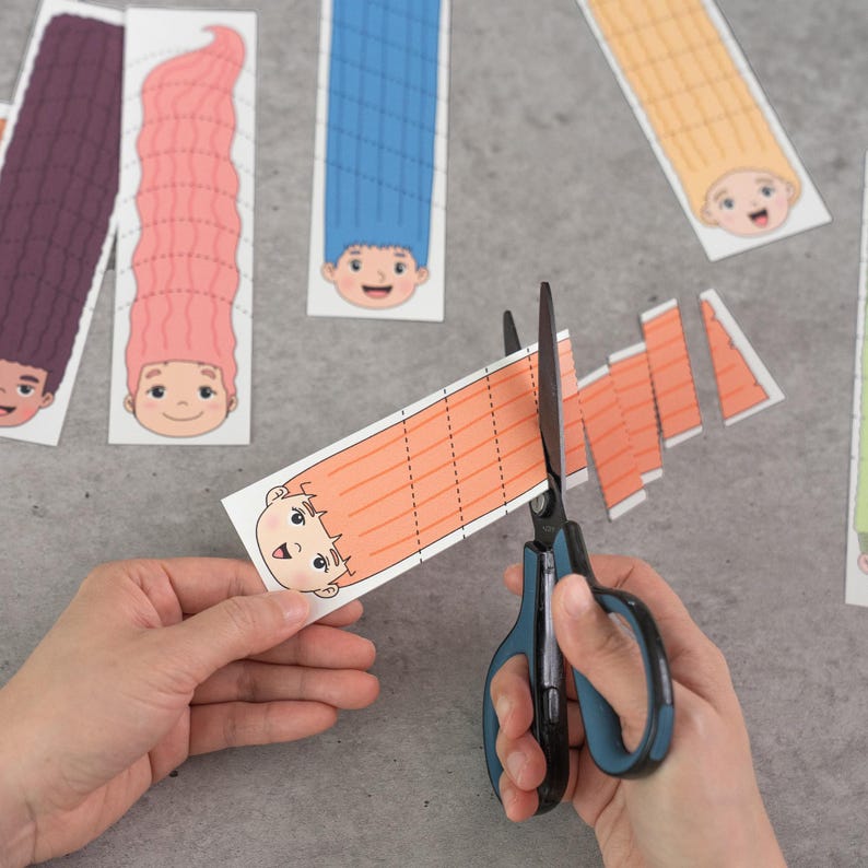 May include: A pair of scissors cutting a strip of orange paper with a cartoon face and long hair. Other similar paper cutouts with different hair colors are scattered around. The image suggests a craft or DIY project.