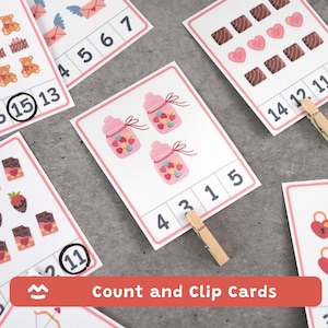 Pode incluir: Cartões educativos de contagem e clip com tema do Dia dos Namorados. Os cartões apresentam ilustrações de ursinhos de pelúcia, chocolates e potes cheios de doces em forma de coração. Os números são exibidos para a prática da contagem. Um prendedor de roupa de madeira é usado para prender a resposta correta.