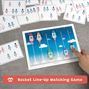 Puede incluir: Un juego de emparejamiento Rocket Line-Up con cohetes de colores en tarjetas y un tablero de juego. El tablero presenta un fondo de cielo azul con nubes blancas y rastros de lanzamiento de cohetes. El juego incluye múltiples tarjetas con patrones de cohetes.