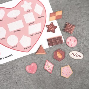 May include: A heart-shaped educational chart with various shapes labeled, including a star, square, and circle. Scattered around the chart are several shape-themed stickers, such as a chocolate bar and a pink heart with white polka dots.